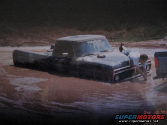 Dark 1977 Ford F-150 stuck in mud, driver's side visible, person reaching near front bumper