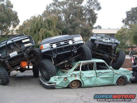 Three monster trucks piled on top of a crushed green sedan