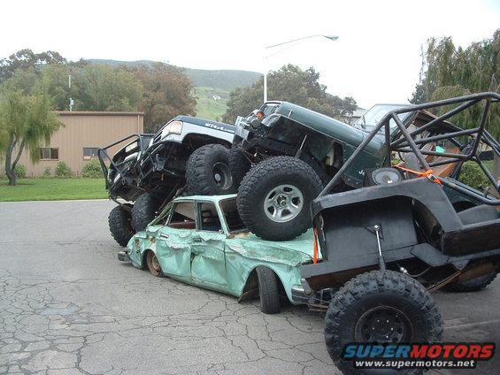 Crushed light blue sedan with multiple off-road vehicles stacked on top