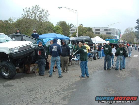 Several people stand around modified off-road vehicles, some under tents, at an outdoor event