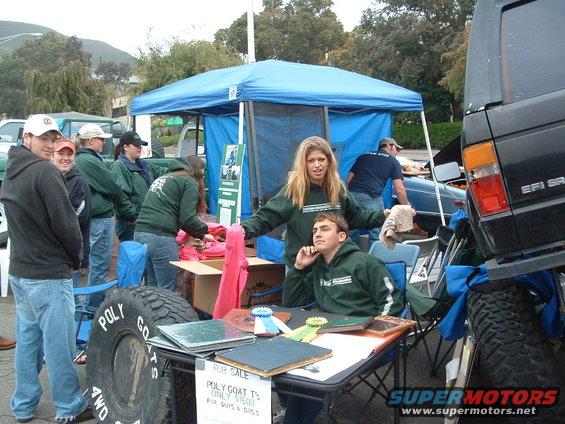 People gathered around a table and tent at an outdoor event, with a large tire and part of a black SUV visible