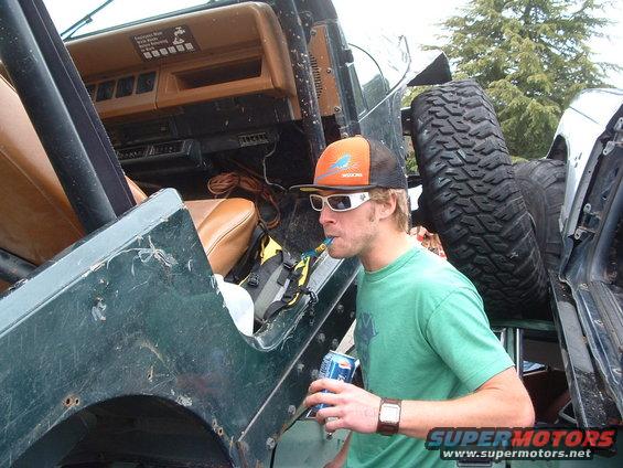 Green Jeep YJ interior with tan seats, man in orange hat drinking from a can, spare tire visible