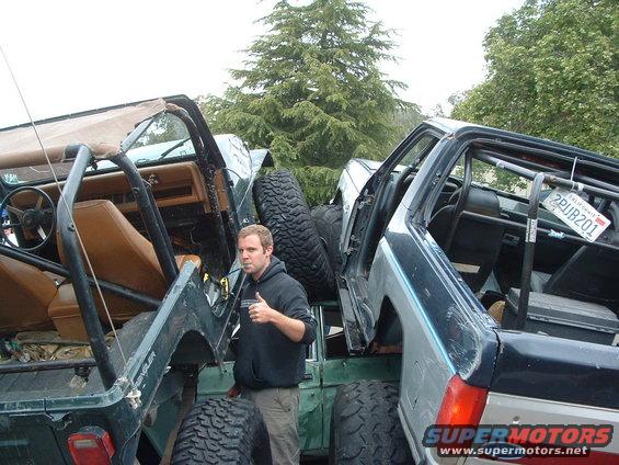 Two Jeep YJs, one green and one dark blue, parked close together. A man stands between them giving a thumbs up