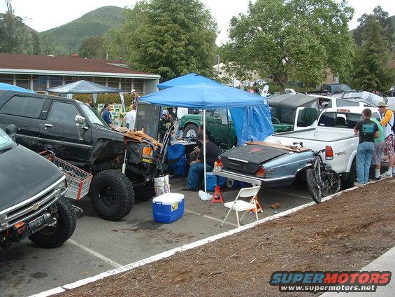 Modified black SUV with large tires, a blue cooler, and a shopping cart, parked next to a blue car on a trailer and a white pickup truck