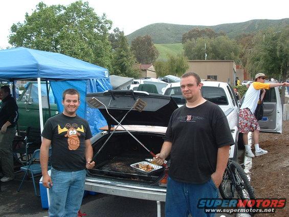 Two men stand by an open car trunk with grilling tools and food. Other vehicles and a tent are visible