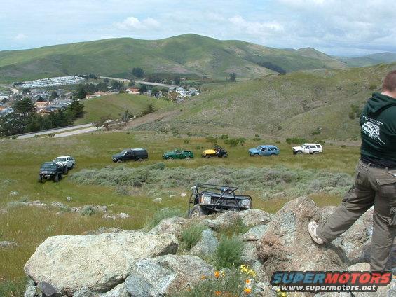 Several off-road vehicles parked and driving on grassy hills with a town in the background