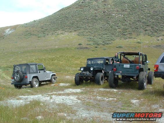 Three Jeeps parked in a grassy field with a hill in the background. One Jeep is silver, and two are dark green