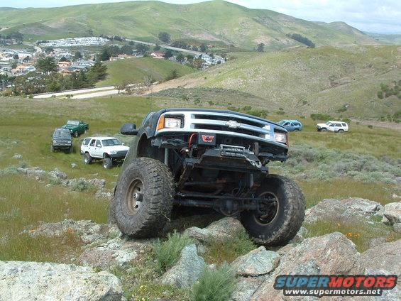 Modified Chevrolet pickup truck with large tires on rocky terrain, other SUVs and trucks in background