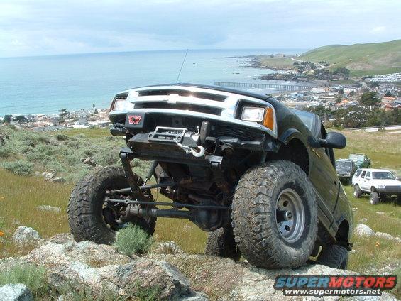 Black SUV with lifted suspension and large tires on rocky terrain, ocean and town in background. Other SUVs visible