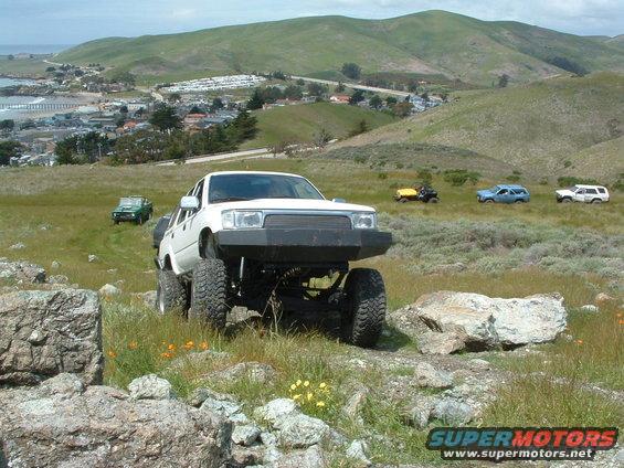 Modified white Toyota pickup truck with large tires and bumper, parked on rocky terrain. Other vehicles visible in background