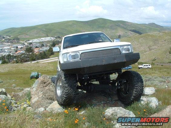 White off-road truck with large tires climbing rocks, with a green Jeep and white SUV in the background