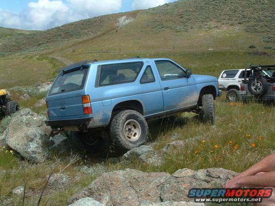 Blue SUV with off-road tires on rocky terrain, rear-left quarter view. Another white SUV and a yellow ATV are visible in the background