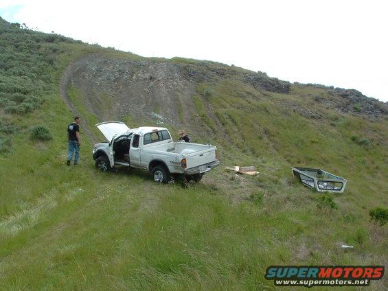 White pickup truck with hood open and door ajar on grassy hillside, removed truck cap nearby