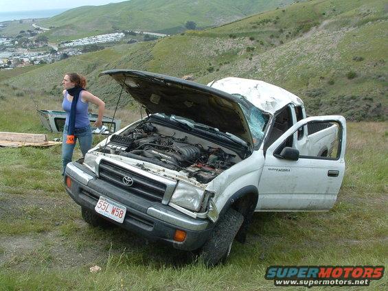 Damaged white Toyota Tacoma pickup truck with hood open and driver's door ajar, parked on a grassy hillside