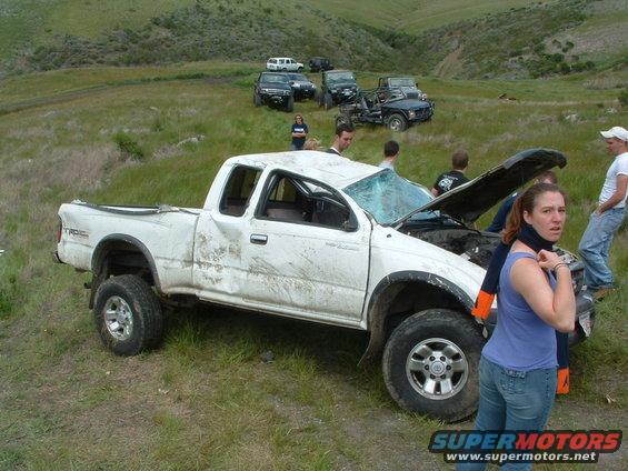 White pickup truck with smashed windshield and open hood, people around it. Other vehicles in background