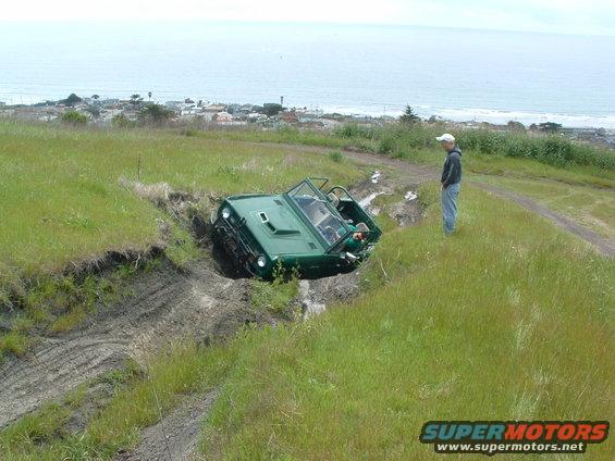 Green off-road vehicle tilted in a muddy ditch with a person standing nearby on a grassy hill overlooking the ocean
