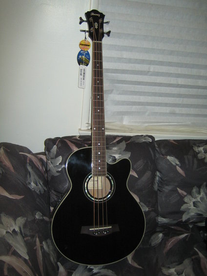 Black acoustic bass guitar with Ibanez logo on headstock, resting on a floral patterned couch