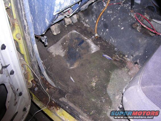 View of the driver's side floorboard and lower door jamb of a dirty vehicle. Shows rust and exposed wiring