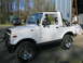 1987 Suzuki Samurai - A white 1987 Suzuki Samurai convertible SUV is parked in front of a metal building with another truck in the background.