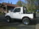 1987 Suzuki Samurai - White 1987 Suzuki Samurai parked outside a wooden house on a sunny day.