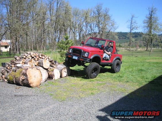 Red 1987 Suzuki Samurai on a grassy field with a pile of logs