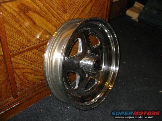 chrome wheel with five spokes, sitting on a dark carpet next to a wooden cabinet