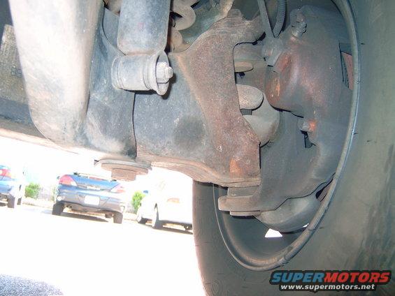 Underside view of a vehicle's front suspension and brake caliper, with other cars visible in the background