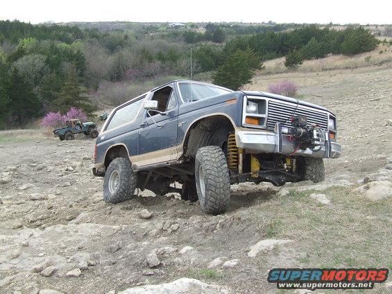 Dark blue Ford Bronco with lifted suspension and large tires driving on a rocky trail. Another green Bronco is visible in the background