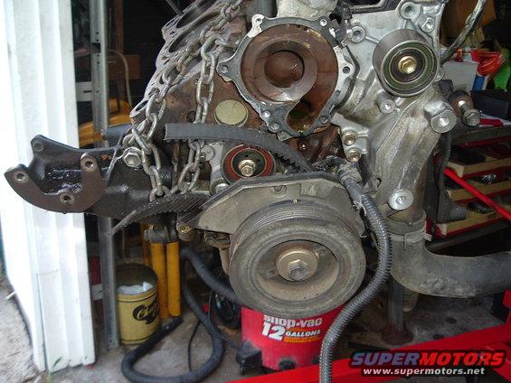 Engine block with timing belt, tensioner pulley, and crankshaft pulley visible. A chain is also wrapped around the block