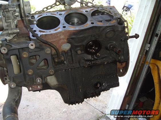 Engine block with oil residue, hanging by chains, with a partial yellow object and metal shelving in the background
