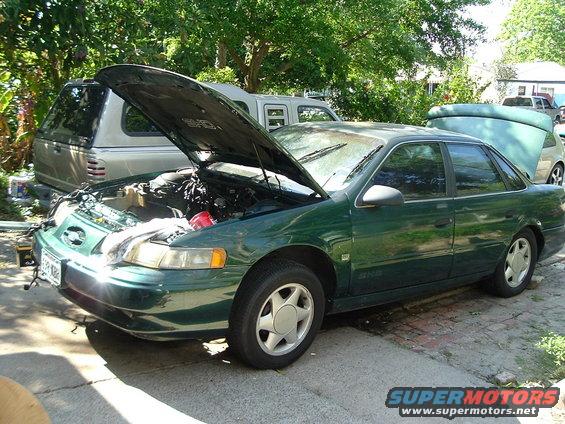 Green sedan, hood up, parked outside next to a silver pickup truck with a cap. Another vehicle is partially visible in the background
