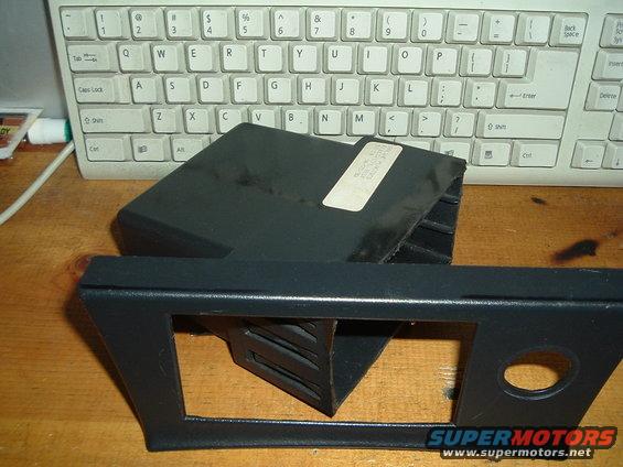 Black plastic car dashboard trim piece and a black electronic module with a label on top, placed on a wooden surface in front of a computer keyboard