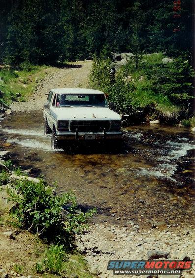 White 1978 Ford Bronco crossing a rocky stream, trees and dirt path in background
