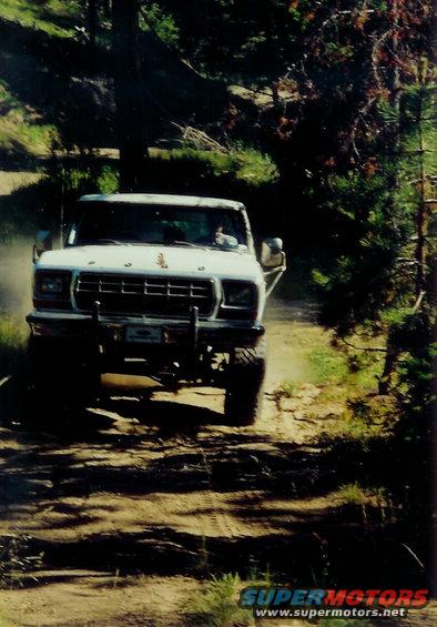White 1979 Ford Bronco driving on a dirt road, front view. Off-road bumper and tires are visible