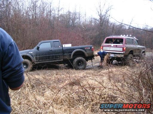 1978 Ford Bronco is stuck in mud and being pulled by a black Ford Ranger. A person is connecting the tow strap