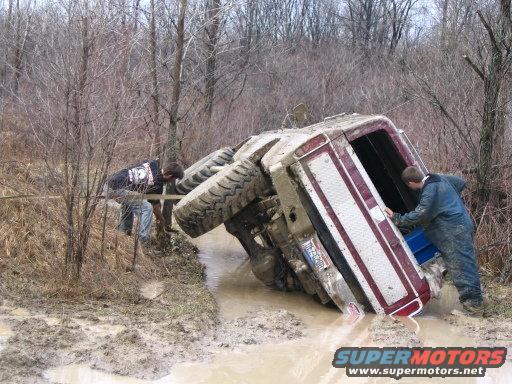 Red and white 1978 Ford Bronco tipped on its side in mud with two people nearby