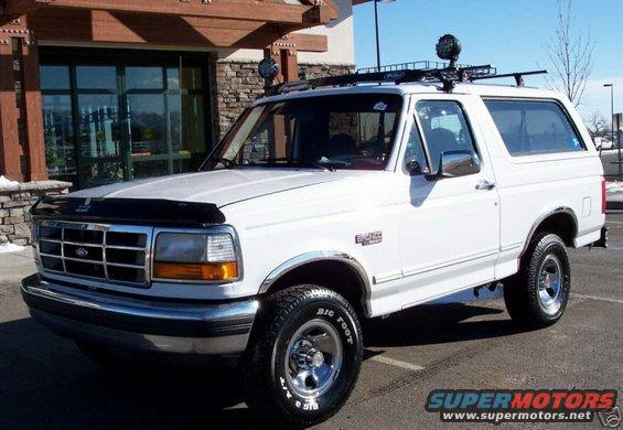 White Ford Bronco with chrome wheels, "Bigfoot" tires, and a roof rack, viewed from the front-right