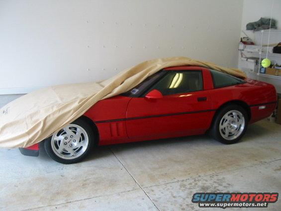 Red sports car, possibly a Corvette, covered with a tan car cover. Side view shows the wheels and part of the body