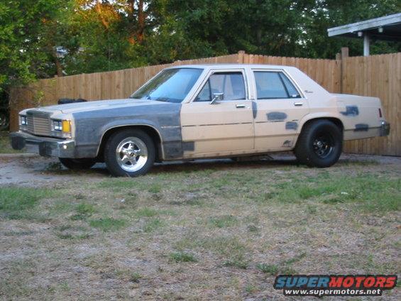 Gray and tan sedan with primer patches, chrome front wheels, black rear wheels, and a wooden fence behind it