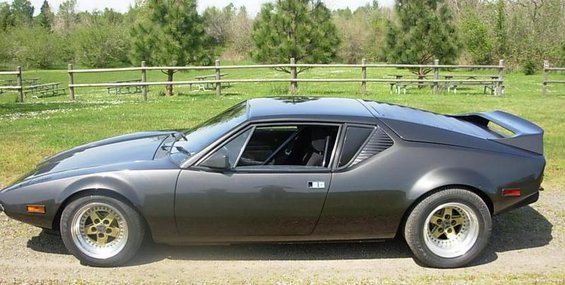 Side view of a dark gray 1972 DeTomaso Pantera with gold-accented wheels parked on a dirt road