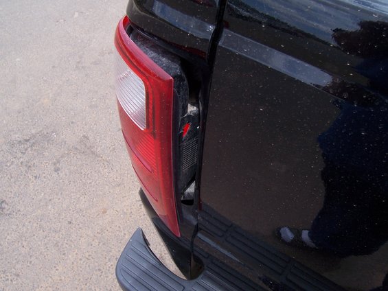 Black pickup truck's damaged rear passenger-side taillight assembly, showing exposed red and clear plastic with a black bumper step