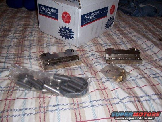 Two metal brake caliper brackets and bags of hardware on a plaid surface, with a Priority Mail box in the background