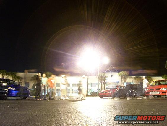 Several cars parked at night with a group of people standing nearby under bright lights