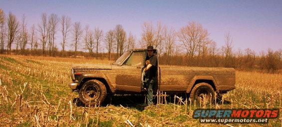 Mud-covered 1970s pickup truck, possibly a Jeep J-10, in a dry field with a person standing by the driver's door