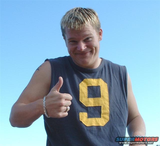Man in a dark blue t-shirt with a yellow number 9, giving a thumbs up gesture. He has blond hair and a tattoo on his left arm. Text "SUPERMOTORS www.supermotors.net" is visible