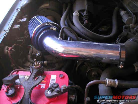 Engine bay showing a blue cone air filter connected to a chrome intake tube, with a red battery in the foreground. SuperMotors.net logo visible