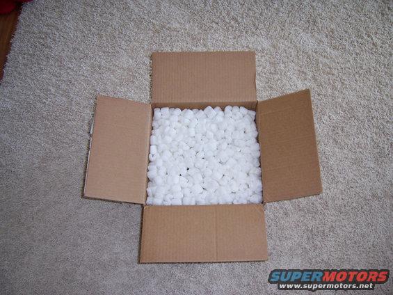 open cardboard box filled with white packing peanuts on a carpeted floor. Supermotors.net logo in corner