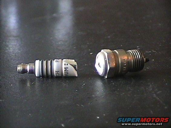 Two spark plugs on a dark surface. One is white with black bands and text, the other is metallic with a white top