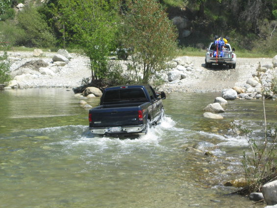 Dark blue pickup truck driving through a river, with a white truck carrying gear behind it