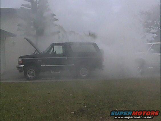 Black 1994 Ford Bronco with hood open, emitting a lot of smoke. Another vehicle is partially visible to the right, also in smoke